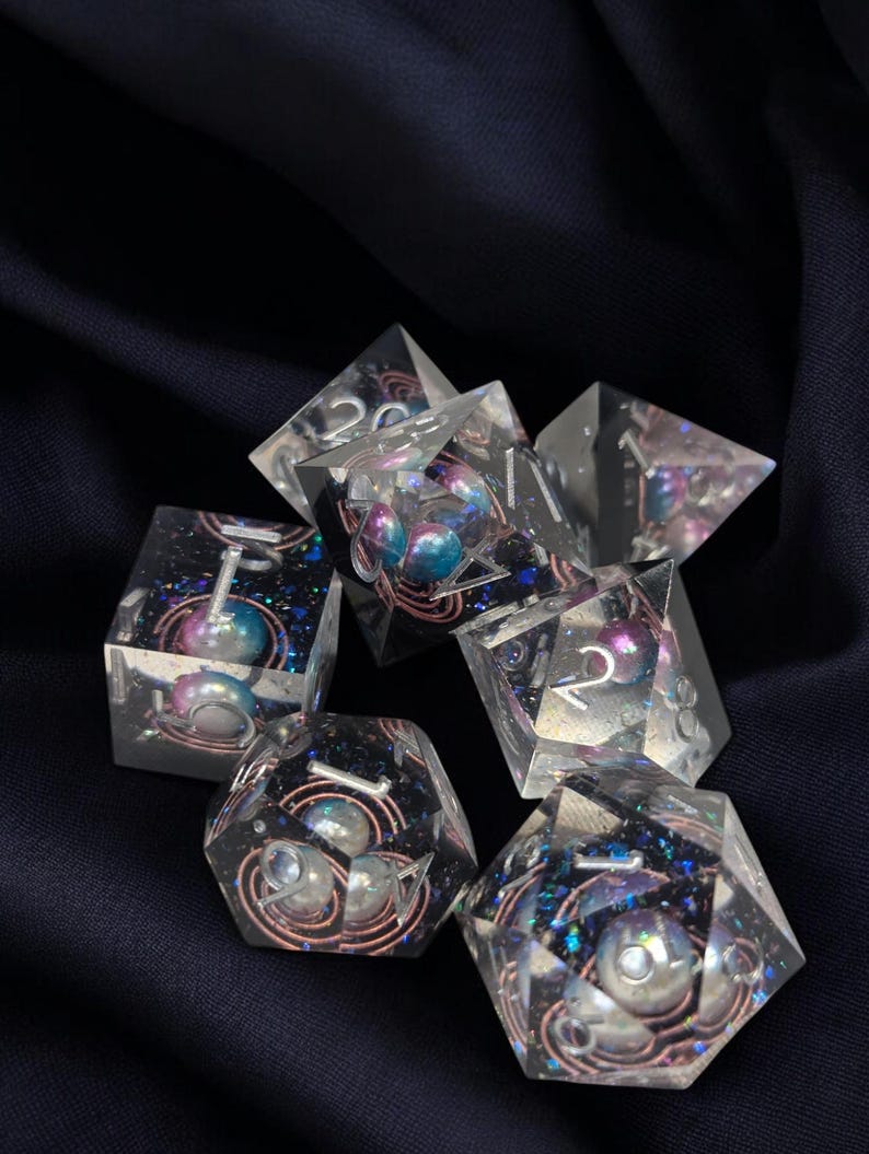 May include: A set of seven clear resin dice with iridescent glitter and embedded pearls. The dice are in various shapes and sizes, with numbers visible on each face.