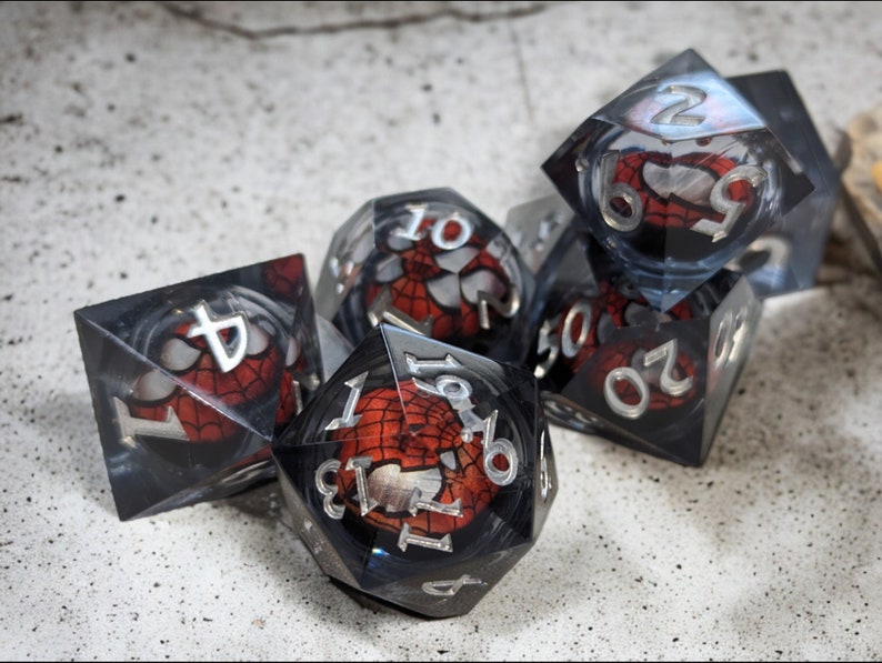 K&ouml;nnte beinhalten: Ein Satz von sechs transparenten W&uuml;rfeln mit einem roten und schwarzen Spider-Man-Design. Die W&uuml;rfel sind von 1 bis 6 nummeriert.