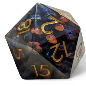 May include: A black and gold d20 dice with a galaxy-themed design. The numbers are in gold and the dice has a clear, iridescent finish.