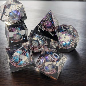 May include: A set of six iridescent dice with a galaxy theme. The dice are clear with embedded glitter and swirls of color. Each die has a different number on each face, from 1 to 6.
