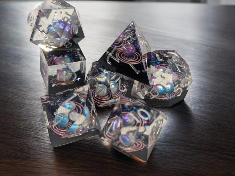 May include: A set of six iridescent dice with a galaxy theme. The dice are clear with embedded glitter and swirls of color. Each die has a different number on each face, from 1 to 6.