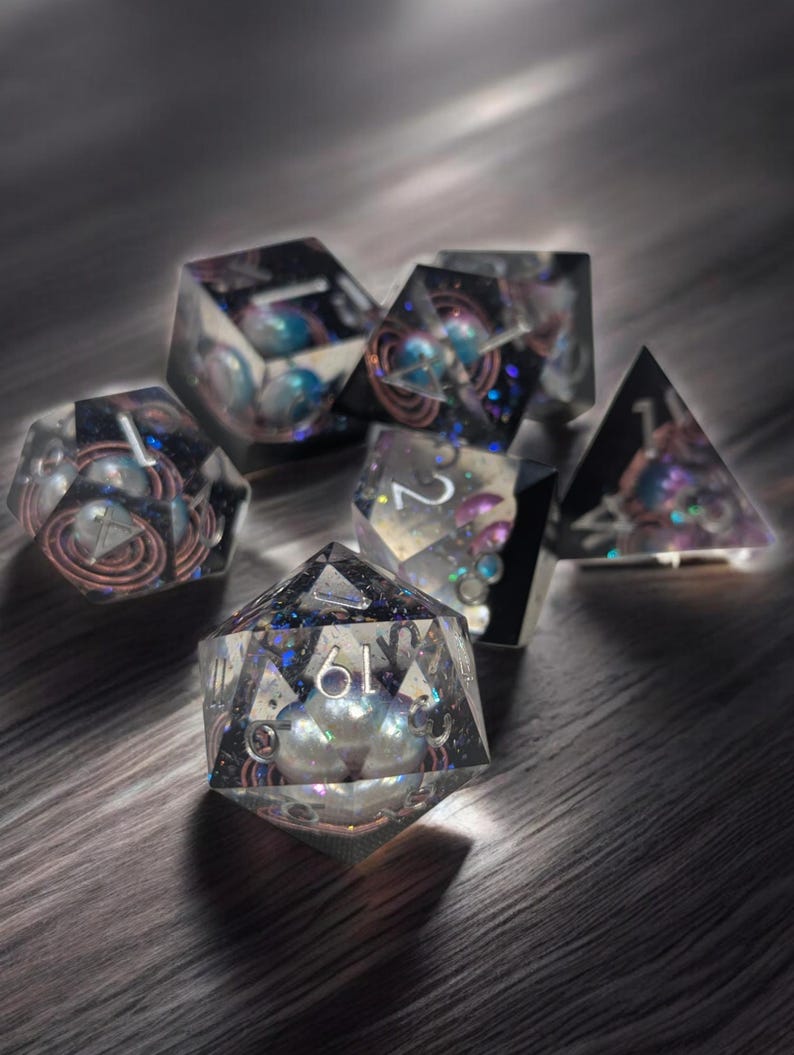 May include: A set of six iridescent dice with a galaxy theme. The dice are made of clear resin with embedded glitter and swirls. The numbers are visible on each face.