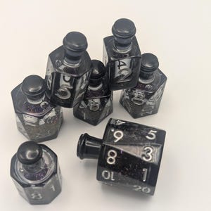 May include: A collection of black, faceted dice bottles and a large, black twenty-sided die. The bottles have black caps and contain glitter. The die has white numbers. The items are arranged on a white surface.