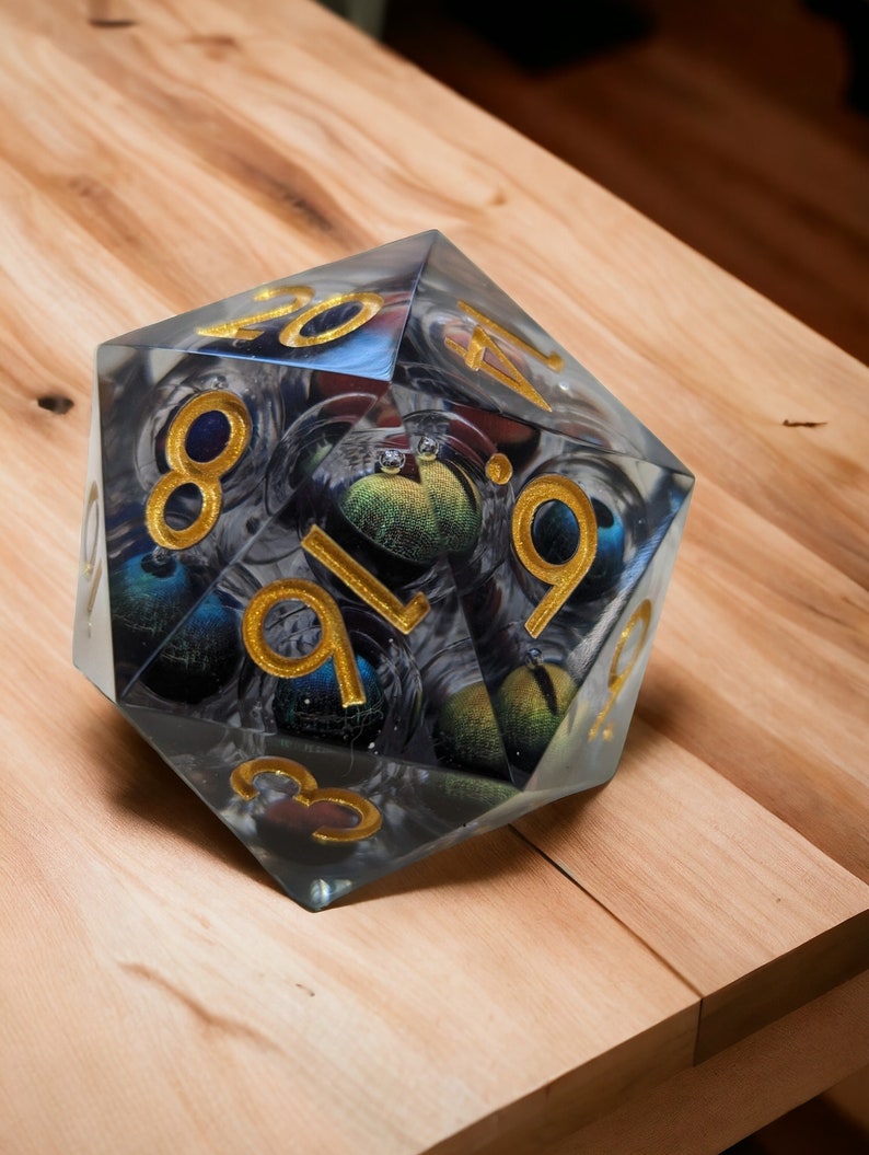 May include: A clear, multi-sided dice with gold numbers and a swirling, galaxy-like pattern inside. The dice is sitting on a wooden surface.