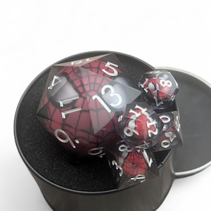 K&ouml;nnte beinhalten: Ein Satz roter und schwarzer W&uuml;rfel mit einem Spider-Man-Design in einem schwarzen runden Beh&auml;lter mit schwarzem Schaumstoff-Innenleben.