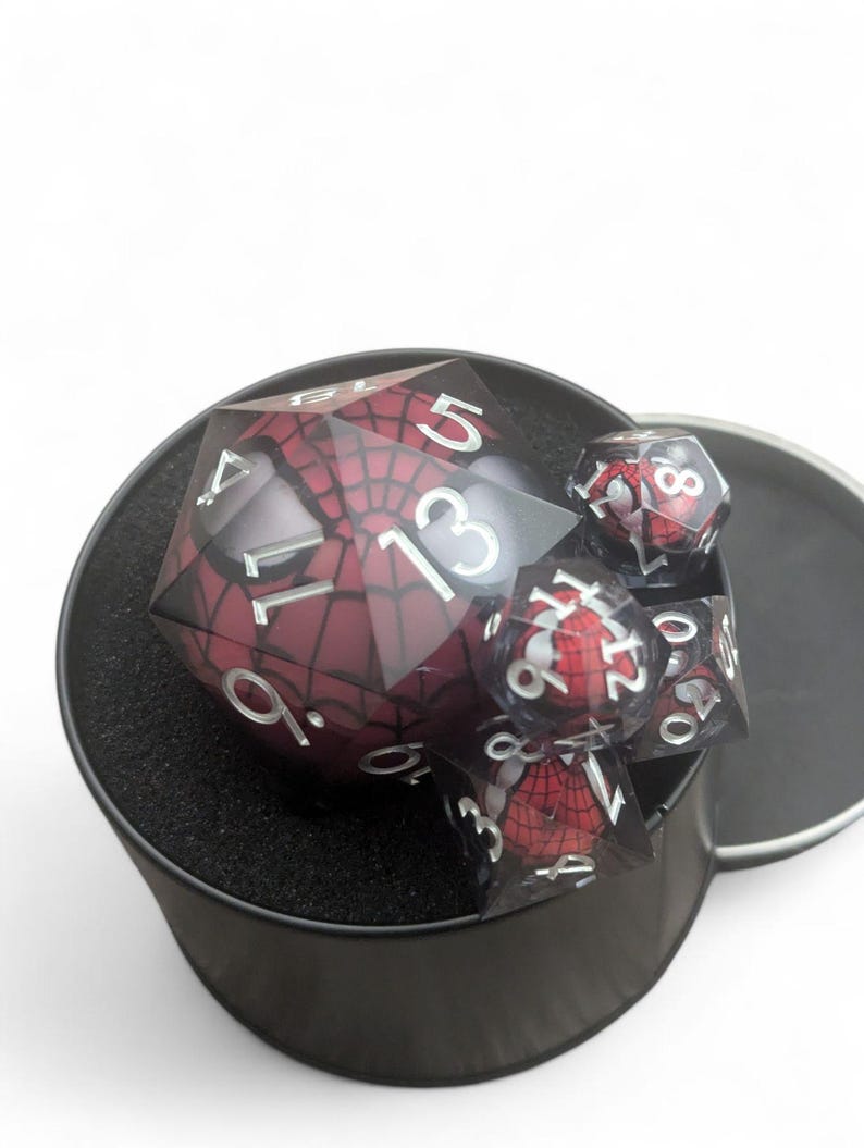 K&ouml;nnte beinhalten: Ein Satz roter und schwarzer W&uuml;rfel mit einem Spider-Man-Design in einem schwarzen runden Beh&auml;lter mit schwarzem Schaumstoff-Innenleben.
