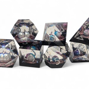 May include: Set of seven iridescent dice with a galaxy theme. Each die has a different number and a silver pearl embedded in the center. The dice are made of clear resin with a black and purple swirl.