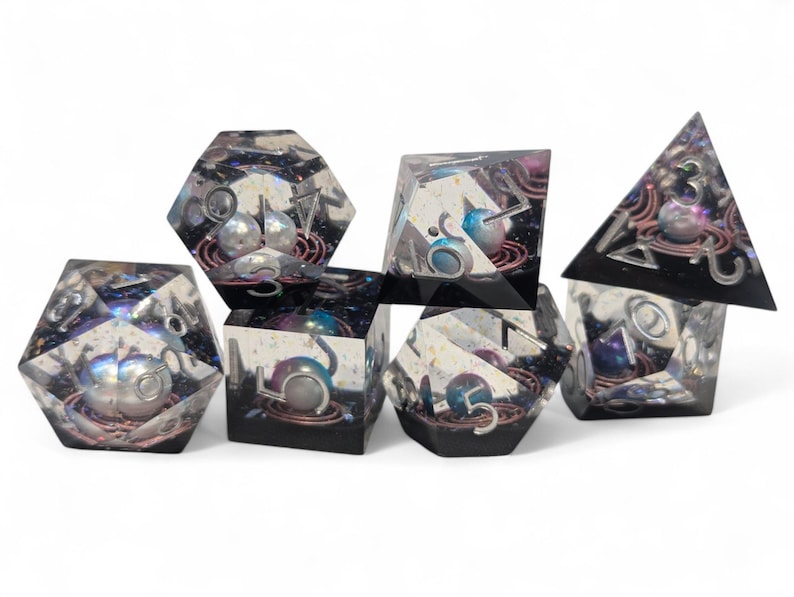 May include: Set of seven iridescent dice with a galaxy theme. Each die has a different number and a silver pearl embedded in the center. The dice are made of clear resin with a black and purple swirl.