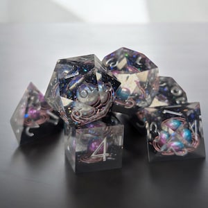 May include: A set of six iridescent dice with a galaxy theme. The dice are clear with embedded glitter and small, colorful beads. The numbers on the dice are visible and easy to read.