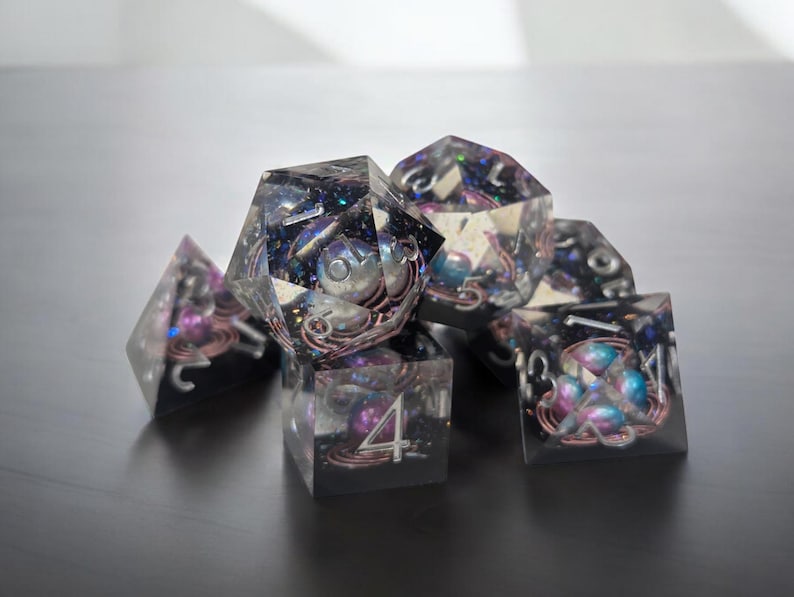May include: A set of six iridescent dice with a galaxy theme. The dice are clear with embedded glitter and small, colorful beads. The numbers on the dice are visible and easy to read.