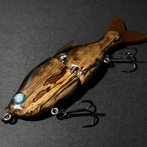 May include: A wooden fishing lure shaped like a fish, featuring a brown and black wood grain pattern. The lure has a clear, glossy finish, a blue eye, and three black fishing hooks. The tail is a translucent brown color.