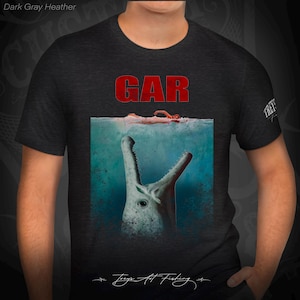 May include: Dark gray heather t-shirt with a graphic of a large, toothy creature emerging from the water with a person's arm and hand visible above the waterline. The text "GAR" is printed in red above the graphic. The text "Treys Art Fishing" is printed in white at the bottom of the graphic.