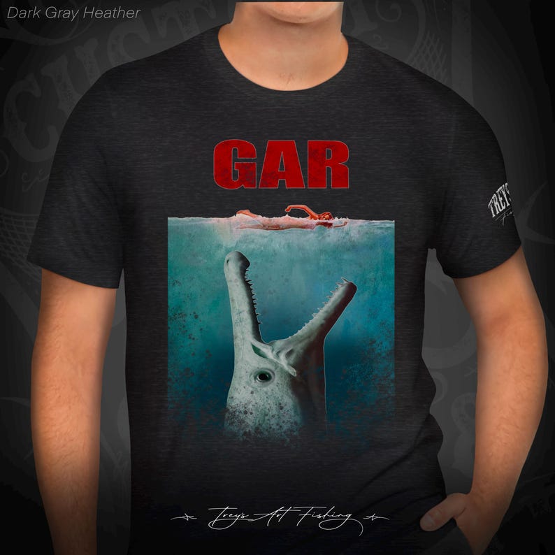 May include: Dark gray heather t-shirt with a graphic of a large, toothy creature emerging from the water with a person's arm and hand visible above the waterline. The text "GAR" is printed in red above the graphic. The text "Treys Art Fishing" is printed in white at the bottom of the graphic.