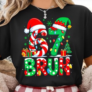 May include: Black t-shirt with a Christmas graphic. The design shows the numbers "67" decorated with candy cane stripes and Christmas lights, and the word "BRUH" in a festive font with lights. Presents and a Christmas tree are also included.