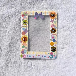 May include: A rectangular photo frame with a yellow and white polka dot border. The frame is decorated with colorful stickers of stars, rainbows, and cartoon faces. A purple bow is attached at the top.