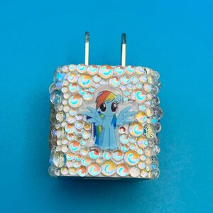 May include: A white charger with a cartoon image of a blue pony with a rainbow mane and tail. The charger is covered in iridescent rhinestones and clear bubble-shaped embellishments. The background is a solid blue.
