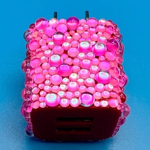 May include: A decorative pink USB charger with a bubbly, iridescent design. The charger has a dark red base with two USB ports and a plug with two prongs. The charger is set against a bright blue background.