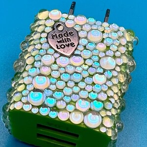 May include: A green and iridescent-covered phone charger with a silver heart charm that reads "Made with Love." The charger is covered in clear, iridescent, and green beads. The background is a solid light blue.