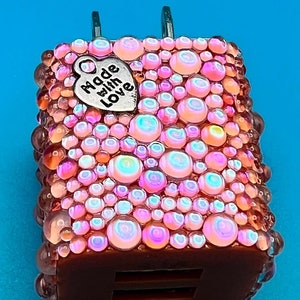 May include: A brown USB charger covered in iridescent pink and orange rhinestones. A small silver heart charm with the words "Made with Love" is attached to the top of the charger. The charger has two metal prongs.
