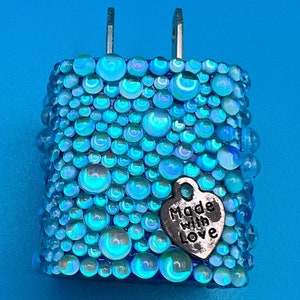 May include: A decorative blue and teal wall plug with a textured surface of iridescent glass beads. A small silver heart charm with the words "Made with Love" hangs from the side. The plug has two metal prongs.