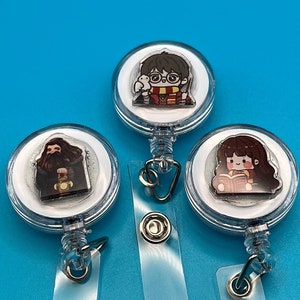 May include: Three clear plastic retractable badge holders with cartoon images of characters. One features a boy with glasses and a scarf, another shows a bearded man, and the third depicts a girl reading a book. The background is a solid blue.
