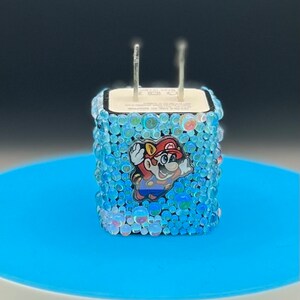 May include: A blue and white charger adapter decorated with iridescent beads. A cartoon image of Mario is featured on the front. The charger has two metal prongs.
