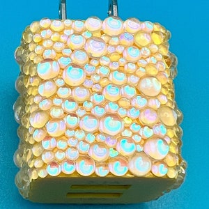 May include: A decorative phone charger with a yellow base and iridescent, bubble-like embellishments. The charger has two silver prongs and a slot for a charging cable. The bubbles reflect a rainbow of colors against a bright blue background.