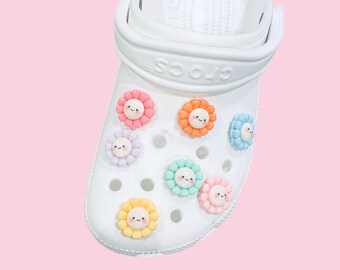 May include: White clogs with colorful flower charms. The charms have smiling faces and are in shades of pink, blue, yellow, and green.