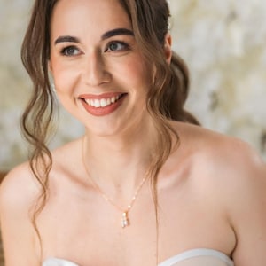Peut inclure: Une mariée portant un haut de corset blanc sans bretelles avec des détails en dentelle florale. Elle sourit et regarde sur le côté. Elle porte un collier en or délicat et un bracelet en perles.