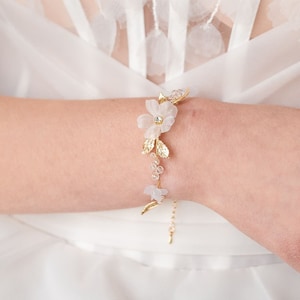 Op de afbeelding: Een delicate gouden armband met witte bloemenaccenten en kristallen versieringen. De armband heeft een ketting met een bloemmotief.