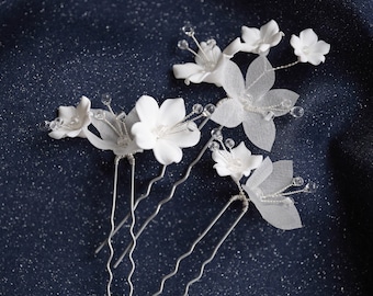 Puede incluir: Cuatro horquillas para el pelo con flores blancas, alambre plateado y detalles de cristal sobre un fondo azul oscuro.