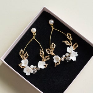 Könnte beinhalten: Ein Paar goldene Ohrringe mit weißen Blumenakzenten und klaren Kristallen, präsentiert in einer weißen Schachtel mit schwarzem Innenraum.