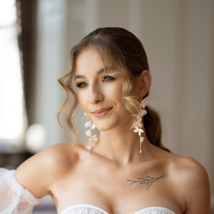 Pode incluir: Uma noiva usando um vestido de noiva branco de ombro caído com um corpete de renda e uma saia de tule. Ela está usando brincos florais delicados e uma pulseira.