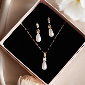 Può includere: Un set di gioielli color oro, composto da collana e orecchini, presentato in una scatola rosa con interno nero. La collana e gli orecchini presentano perle a goccia e dettagli scintillanti. Il set è esposto su una superficie satinata.
