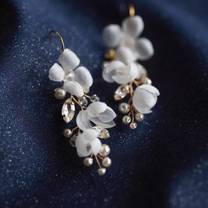 Op de afbeelding: Een paar delicate witte bloemoorbellen met gouden draad en parel accenten. De oorbellen zijn op een donkerblauwe achtergrond.