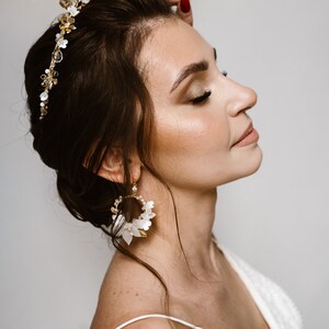 May include: A woman wearing a white dress with a gold and white floral crown and matching earrings. The crown is made of small flowers and pearls, and the earrings are large and circular with white flowers and gold accents.