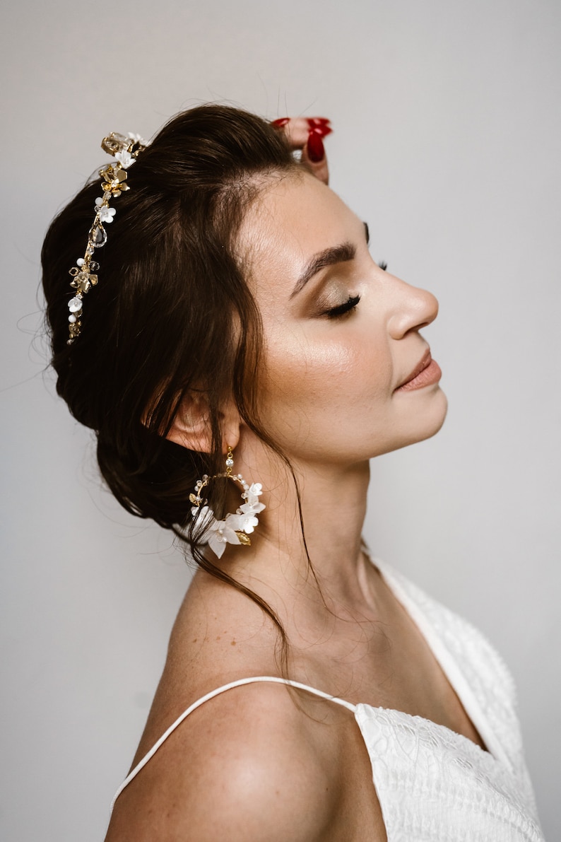 May include: A woman wearing a white dress with a gold and white floral crown and matching earrings. The crown is made of small flowers and pearls, and the earrings are large and circular with white flowers and gold accents.