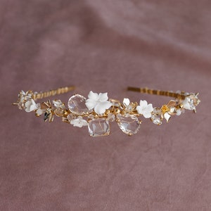 May include: A gold headband with white ceramic flowers and clear crystals. The headband is decorated with small gold beads and leaves.