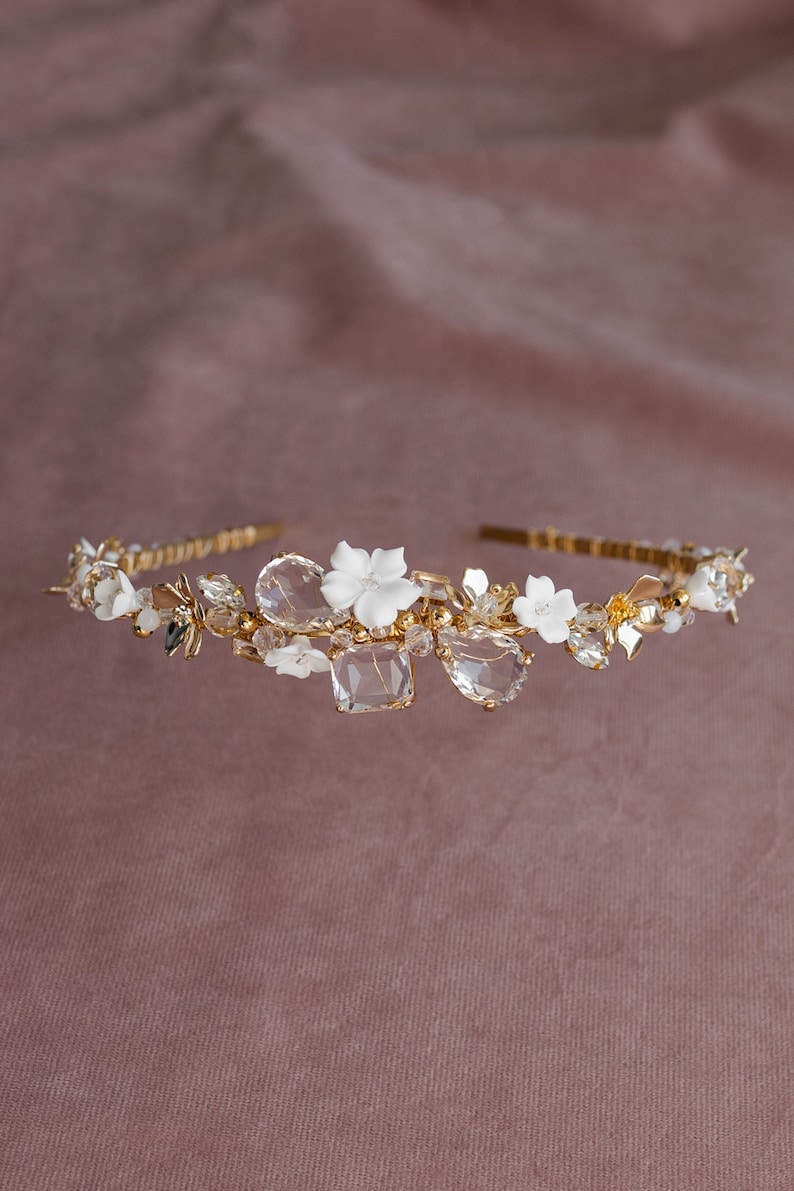 May include: A gold headband with white ceramic flowers and clear crystals. The headband is decorated with small gold beads and leaves.