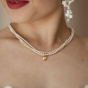 May include: A delicate pearl necklace with a gold heart pendant. The necklace is composed of three strands of small, round pearls. The image also shows a pair of white floral earrings and red lipstick.