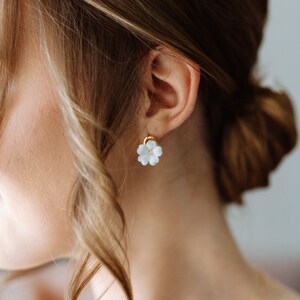 Op de afbeelding: Close-up van een oor versierd met een witte, bloemvormige oorbel met een gouden pin. De persoon heeft lichtbruin haar dat in een knot is gestyled. Een gouden ketting is zichtbaar.