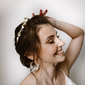 May include: A bride with a white floral crown and matching earrings smiles while holding her hair up. She is wearing a white lace dress.