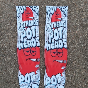 May include: Pair of novelty socks with a red heart-shaped face smoking a cigarette. The socks have the words "POTHEADS" and "POT HEADS" in white, outlined in black, with a cloud design.