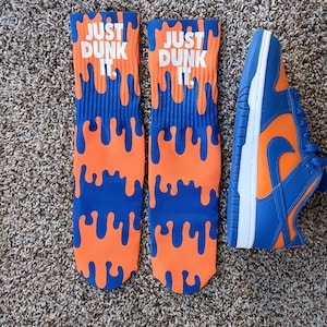 Puede incluir: Un par de calcetines azules y naranjas con el texto "Just Dunk It" impreso en ellos. Los calcetines tienen un diseño de goteo. También se muestra una zapatilla de baloncesto azul y naranja.