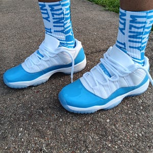 May include: White and light blue sneakers with matching socks. The sneakers feature a glossy light blue toe cap and sole, with white laces and a white upper. The socks are light blue with the word "WZNTY" in white.