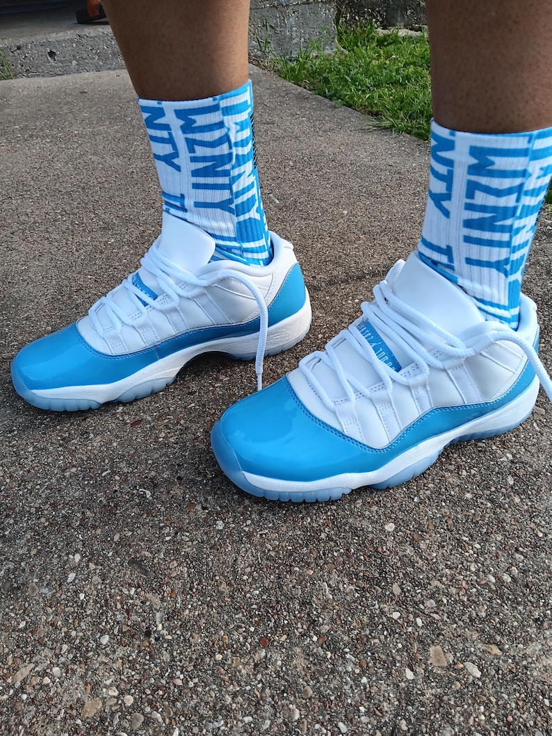 May include: White and light blue sneakers with matching socks. The sneakers feature a glossy light blue toe cap and sole, with white laces and a white upper. The socks are light blue with the word "WZNTY" in white.