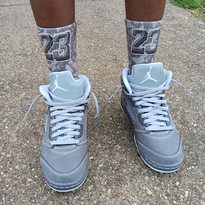 Może przedstawiać: Szare i białe buty sportowe z pasującymi szarymi skarpetami. Skarpety mają numer "23" w ciemnoszarej czcionce. Buty mają białe sznurowadła i kultowe logo Jumpman na języku.