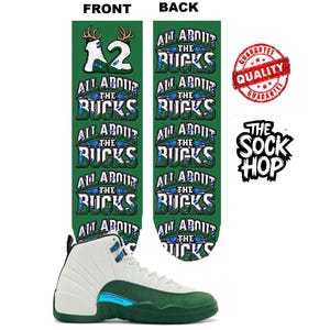 Puede incluir: Calcetines verdes con el texto ALL ABOUT THE BUCKS en un patrón repetitivo, con un gráfico del número 12 y astas de ciervo en la parte superior. Se muestra una zapatilla alta blanca y verde debajo, junto a un sello rojo de garantía.