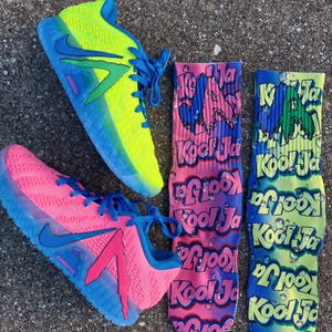 May include: A pair of neon yellow and pink athletic shoes with blue accents and laces are displayed next to two pairs of Kool Ja socks. The socks are pink, blue, and green with the Kool Ja logo.