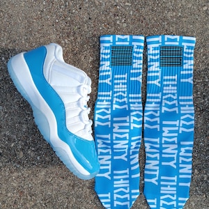 May include: A white and light blue trainer with a clear sole is next to two pairs of blue socks. The socks have the words "TWENTY THREE" in white. The trainer has a shiny light blue toe.
